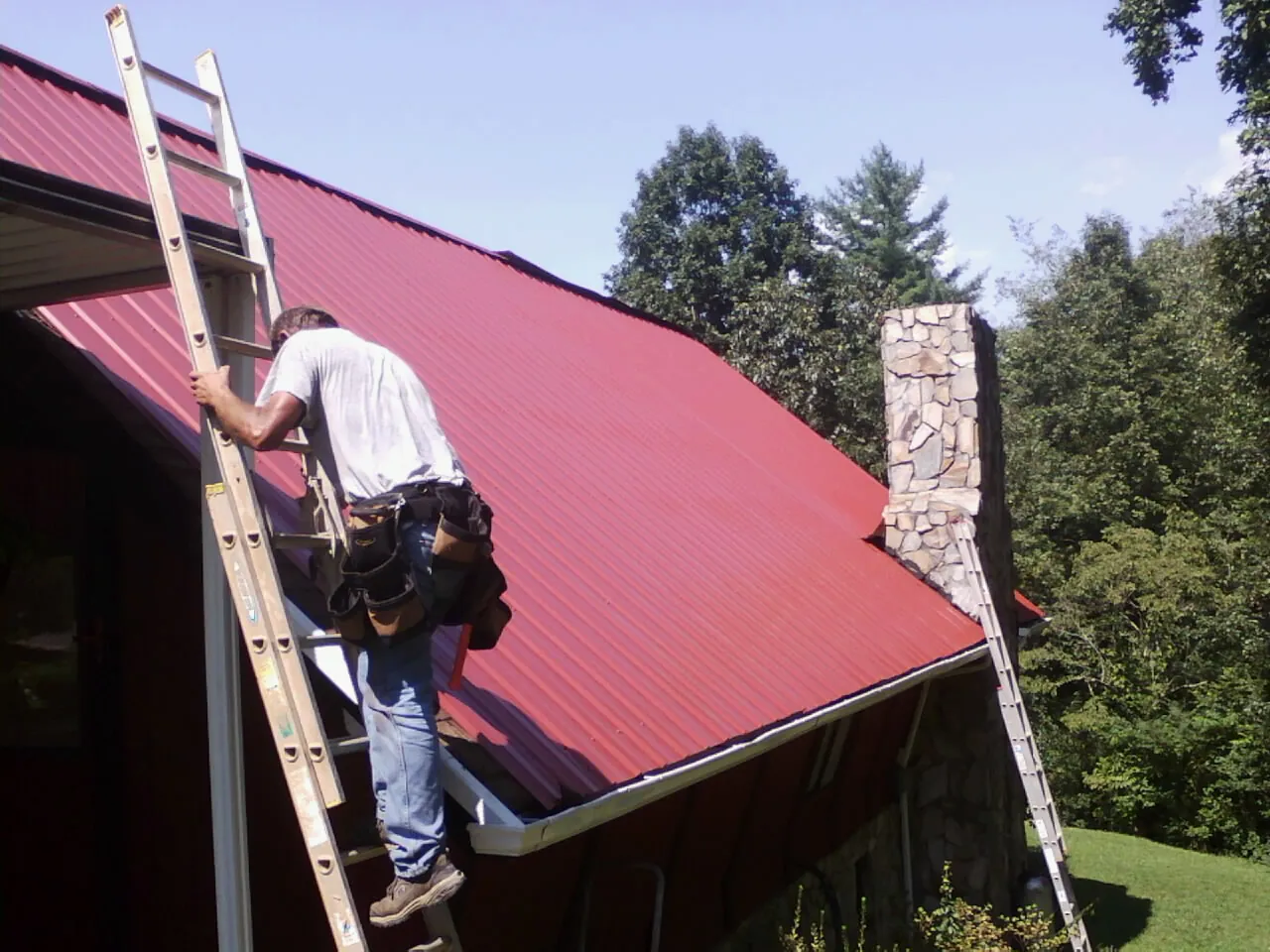 Roofer on a ladder beside a red metal roof and stone chimney — Soffit & Fascia Repair by Roofing Brothers in Cedar Mountain